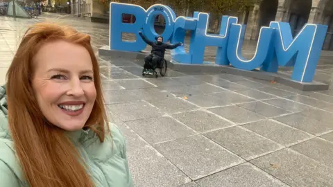 Anna Thomas A selfie of a woman with long red hair, wearing a teal puffer jacket and smiling at the camera. Behind her are large blue letters spelling BOCHUM, and in front of them is a man, dressed in all black and sat in a wheelchair with his arms up in the air.