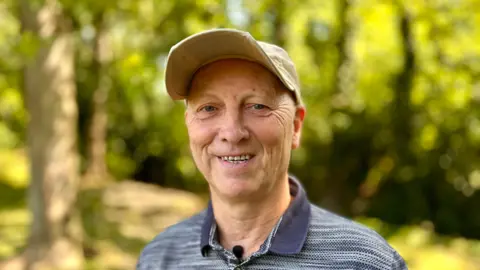 Martin Giles/BBC Mike Wilson stares at the camera with a smile. He wears a beige baseball cap and a collared navy t-shirt. There is a blurred background of trees.
