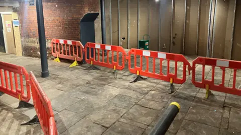 Orange plastic barriers create a passageway along the ground floor of the Meadow Mill apartment block. The barriers can be seen placed a long a stone-paved floor, with parts of the walls stripped back. 