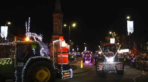 Illuminated tractor with Santa statue on the back in a convoy passinf Banbury Cross at night