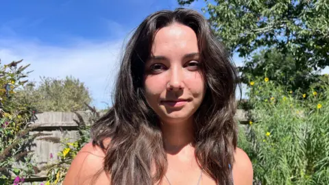 Nicola Haseler/BBC A young woman with long dark brown hair looking into the camera. She is standing in a garden with a wooden fence behind her. There is a tree and foliage in the background.