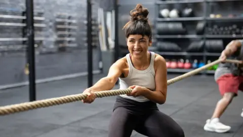 A woman smiles, with her hair in messy top knot, playing tug of war at a cross training gym.