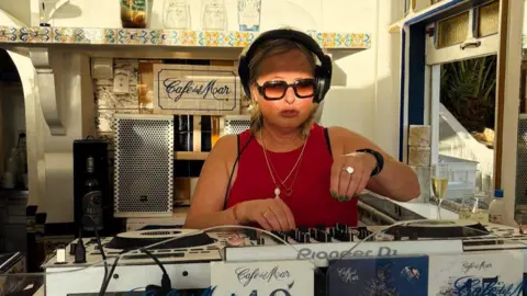 Sally Rodgers Sally Rodgers is wearing an armless red top and wearing sunglasses at a DJ desk.