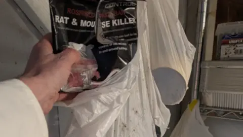 Oxford City Council A white plastic bag is hanging from something out of shot. A hand is pulling a black package out of the bag. The package is open, with a split through its centre, and reads "rat and mouse killer" in white letters. 