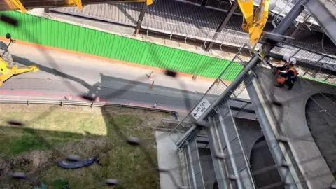 Ross Harvey A worker wearing an orange hi-vis jacket is assessing the damage on an upper level of the car park. There is debris on the floor. A metal barrier is hanging off the end of the car park where the worker is.