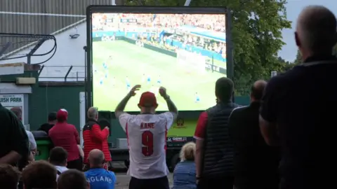 Yeovil Town Football Club A man wearing a Harry Kane England football shirt stands with his arms above bhis head as he and other fans watches the England game on a big outdoor screen.