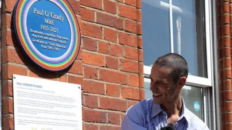 Mainly blue plaque on wall being unveiled by smiling man who is husband of Paul O'Grady