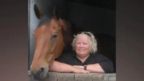 Retired riding instructor Wendy Buckney is stood in a stable next to a horse. She has her arms crossed and has sunglasses atop her blonde hair. She is wearing a black top.