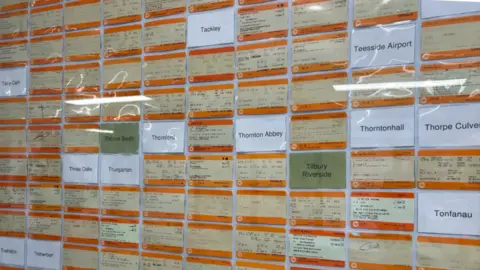 Kate Bradbrook/BBC A wall of bright orange rail tickets, with white pieces of paper with the names of railway stations that the collection does not have tickets for yet