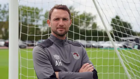 Alex Hughes posing for a picture while stood in a goal wearing a Grimsby Town tracksuit