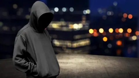 Man wearing black hoodie with face shielded infront of city skyline 