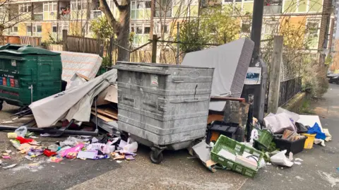Two large bins with lids overflow with rubbish, including two mattresses, in common areas on the estate.