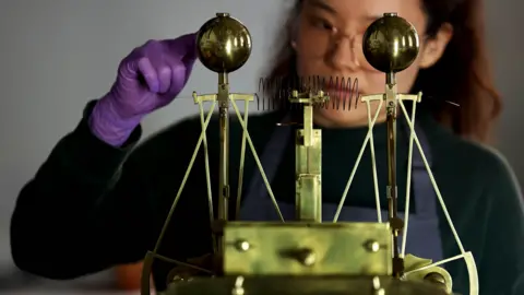 A woman, slightly out of focus, wears purple gloves and a blue apron as she adjusts a large, golden 18th Century timepiece. She is wearing glasses and focusing intently. The timepiece has a rectangular base and two raised arms, each topped with a metallic globe.