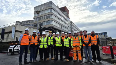 Paul Moseley/BBC A row of men and one woman dressed in hard hits and high-vis clothing stand in front of Anglia Square.