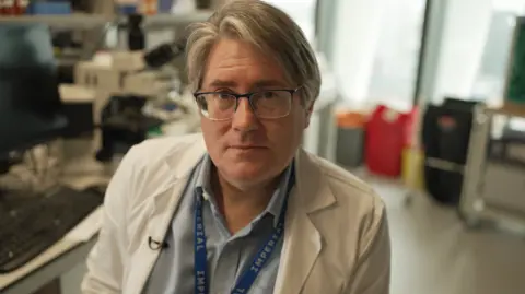 Dr. Ian Mudway en una bata de laboratorio blanco en su universidad. Está mirando la cámara, tiene el pelo gris y lleva un par de gafas. 