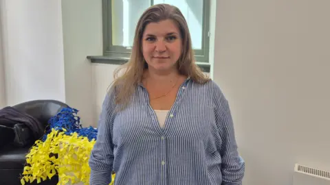 Oksana Khvostenko - a woman with shoulder length blonde hair, wearing a blue and white striped top and standing in a room indoors. 