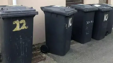 BBC Five black wheelie bins with numbers stand in front of a brown wall. There are numbers painted on a few of the bins.