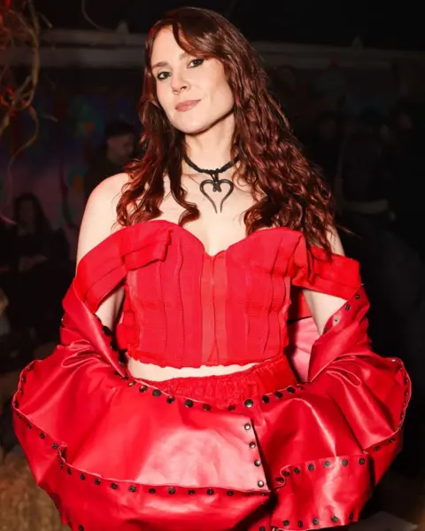 Dave Benett/Getty Images Kate Nash stands smiling at the camera. She is wearing a red top with long sleeves and a heart-shaped necklace