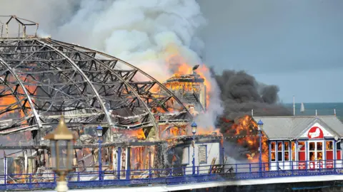 EPA Fire engulfs part of Eastbourne Pier, with bright orange flames tearing through wooden structures and a curved metal framework collapsing and blackening. Thick grey smoke billows into the sky, partially obscuring the sea and coastline. Sections of the building are visibly burned out, while blue railings and lampposts remain in the foreground.