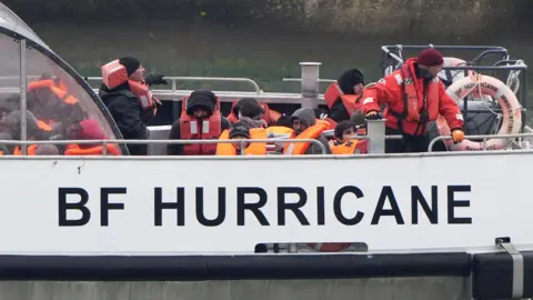 PA Media Eleven people wearing outdoor clothing and orange lifejackets sit on the top deck of a UK Border Force vessel as it sails in to Ramsgate.