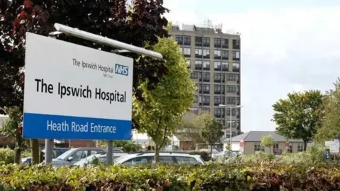 An Ipswich Hospital sign in black writing on a white background. A blue band at the bottom has "Heath Road Entrance" in black writing. There are cars parked behind the sign, which is set into a hedge. There are trees in the car park. A large concrete building of around a dozen stories is beyond the car park.