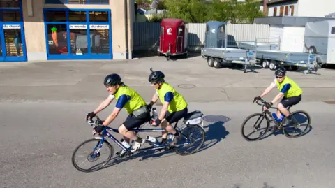 bbc Three male cyclists, two on a tandem, cycle on a town centre road in the sunshine