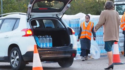 A person wearing an orange hi-vis jacket loading water into the boot of a car