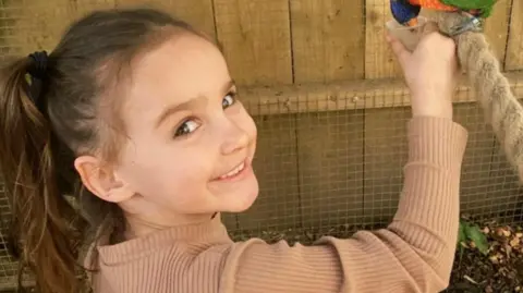 Family handout Aria Thorpe. She is a young child with long, dark hair tied up in a ponytail. She is pictured feeding a colourful bird and is looking over her shoulder to smile at the camera.