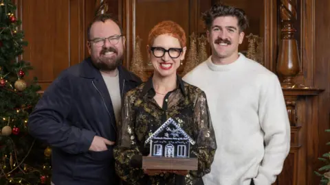 The three judges of Scottish Home of the Year, Anna Campbell-Jones, Banjo Beale and Danny Campbell, stand smiling at the camera. Anna, who is in the middle, is holding the Scottish Christmas Home of the Year trophy, which is shaped like a gingerbread house.