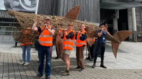 Several people wearing orange hi-vis jackets hold up the Basket Shark outside the National Maritime Museum in Falmouth.