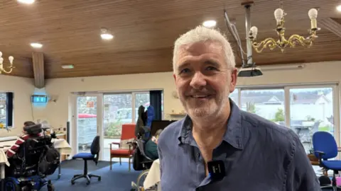 Chairman Rob Shepherd smiling at the camera. He is in a room at the home with floor length windows and some of the residents behind him.