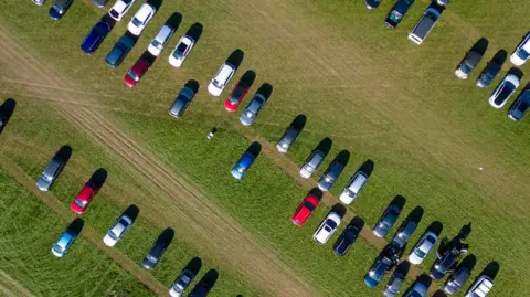 Getty Aerial view of vehicles parked in a green field. The cars are red, blue, black and white.