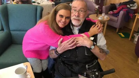 Rachel Davies Rachel Davies is seen smiling at the camera with her arms around her father, Howard Ryder. She is wearing a bright pink jumper and has long, light brown hair. Mr Ryder is hugging his daughter and is wearing spectacles and a checked shirt and jacket. He is sitting in a mobility scooter