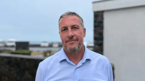 Deputy Andy Cameron, stood smiling with pursed lips. He has grey hair and facial hair and is in his 50s. He is wearing a blue shirt and in the background is a blurred road and grey sky. 