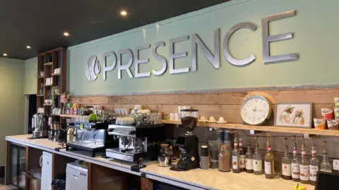 The back of a coffee bar serving area, with bottles of flavoured syrups and a clock as well as coffee machines and cups. There is metal lettering on the wall which reads "Presence".