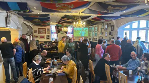 Kitchener's Guest Home At least 30 men and women in a dining room, queuing for food or sitting at tables, eating and talking. The ceiling of the room is swathed in colourful regimental flags. 25.01.25