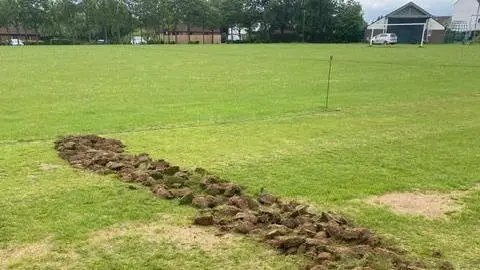 Westcroft Cricket Club A cricket pitch with a long, uneven trench of freshly dug turf running diagonally across the ground. The disturbed soil forms a rough line of clumps, contrasting with the otherwise green and well-maintained grass. In the background, there are houses, parked cars, and a line of trees under an overcast sky. Faint white and blue field markings are visible.