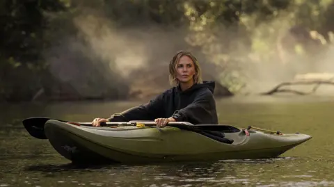 Netflix Theron sat in a green canoe in a grey hoodie. Her blonde hair is shoulder-length