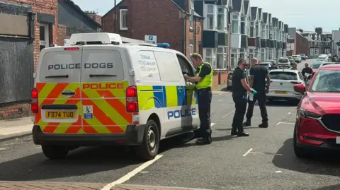 Jim Scott/BBC A white police van with a orange and yellow line pattern on the back doors. At the top of the doors it reads 'Police dogs' in blue lettering. There is an officer in a hi-vis jacket leaning into the window, talking to the driver. There are two other officers dressed in all black in front of them. The white Volkswagon is ahead of them. Cars line the street which is made up of red brick terraced houses.