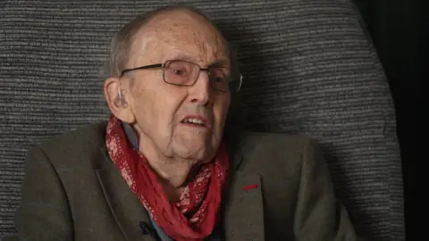 Cecil Newton, an elderly white male wearing thing black framed glasses and a hearing aid, with a red cravat and green tweed blazer sitting in his armchair
