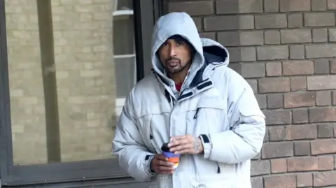 A man wearing a grey hoodie and a grey jacket. He is holding a coffee cup.