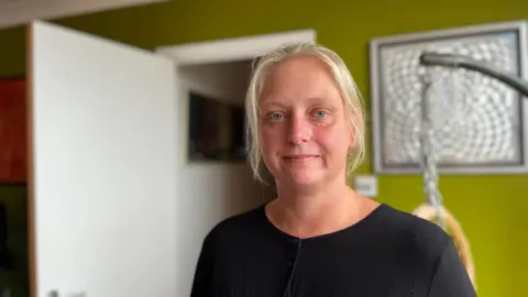 George Carden/BBC Angela Hersee standing in a living room with green paint on the walls. She's wearing a black top and has blonde hair