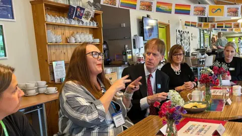 A group of people are gathered around a wooden table in a a café area. The table is decorated with bright, fresh flowers and plates of cookies, suggesting a friendly, social gathering. In the background, shelves hold neatly arranged cups and saucers, and colourful banners celebrating Pride Month hang from the ceiling. The people are engaged in conversation, with the minister, who is listening intently.