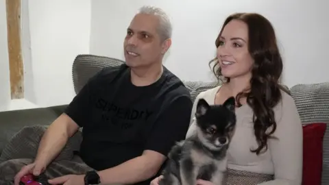 Shaun Whitmore/BBC Nick Samiotis relaxes on the sofa at his home near Watton in Norfolk, 10 days after surgery. He is playing on a games console and both he and his daugther Tabitha, look at the computer screen. She has a small brown and cream dog sitting on her lap. She is wearing a cream top and has brown, long curled hair. He has a black t-shirt on and a black wrist watch.
