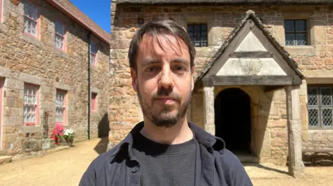 Ben Spink stands centered in a sunlit stone courtyard, facing the camera, wearing a dark shirt and jacket. Behind him are rustic buildings made of warm-toned stone, including one with pink-trimmed windows on the left and another with a thatched roof and a small wooden porch on the right. Bright blue sky and a pot of red and white flowers add colour to the scene.