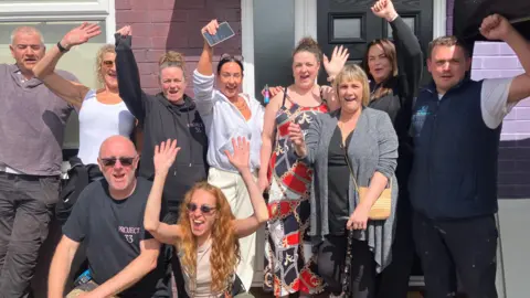 Ten people hold their arms aloft as they celebrate outside the newly-renovated home