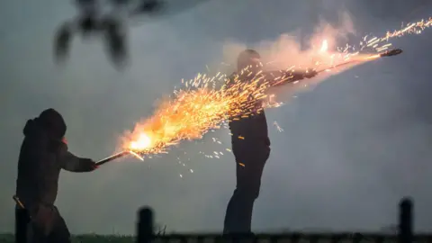 Tony Marsh Two masked youths weaing black trousers and hoodies set off fireworks from their hands, sparks fly and the rockets launch in a smokey night time scene