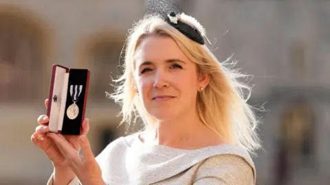 Katherine Goodwin is seen posing for a picture holding her King's Medal. She is wearing a beige dress and black fascinator.
