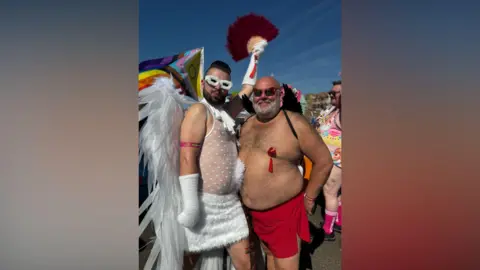 Two barely clothed men. One is topless. The other is dressed as a white angel.