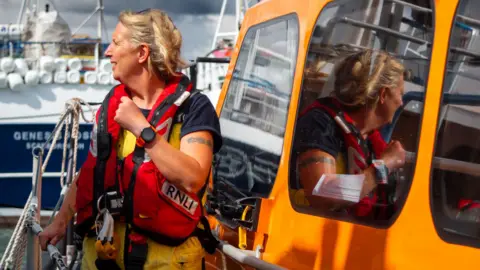 RNLI/Mike Milner Sarah Berrey is standing next to an orange RNLI lifeboat, looking into the distance while wearing her lifeboat safety uniform and red life vest 